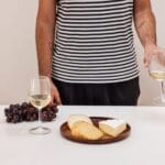 Osoba degustuje białe wino przy półmisku serów i winogron